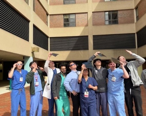 A group of residents wearing scrubs stands outdoors observing the solar eclipse using filters and glasses